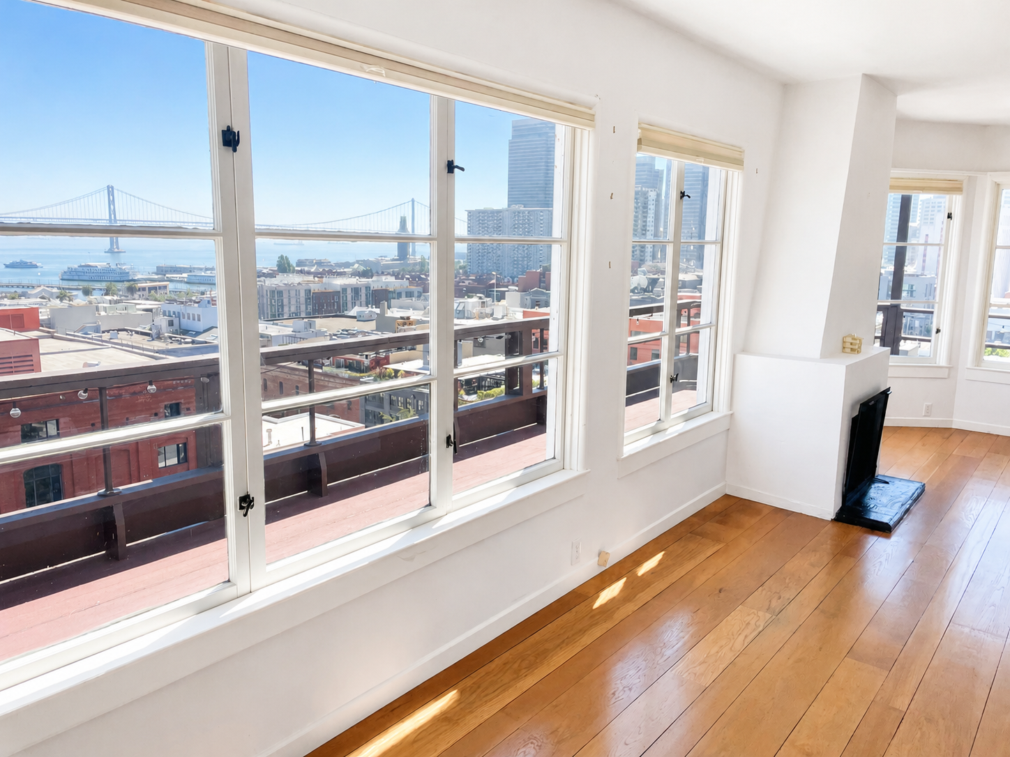 Living room with large windows overlooking a city skyline and water, hardwood floors, and a small fireplace in the corner.
