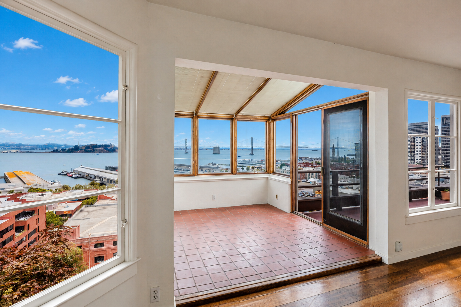 Room with large windows offering a panoramic view of the water, bridge, and city skyline, with a small enclosed balcony or sunroom with terracotta tile flooring.