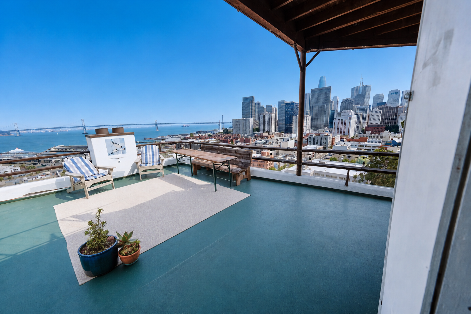 Rooftop balcony overlooking San Francisco skyline, with patio chairs, a table, potted plants, and a view of the bay and bridge.