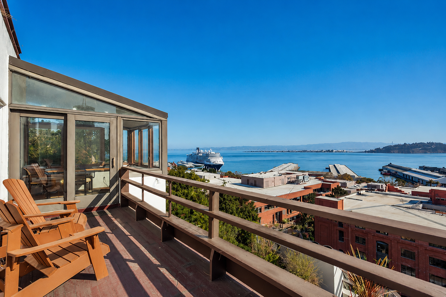 Balcony with wooden chairs overlooking a harbor with a cruise ship and a ferry under a clear blue sky.