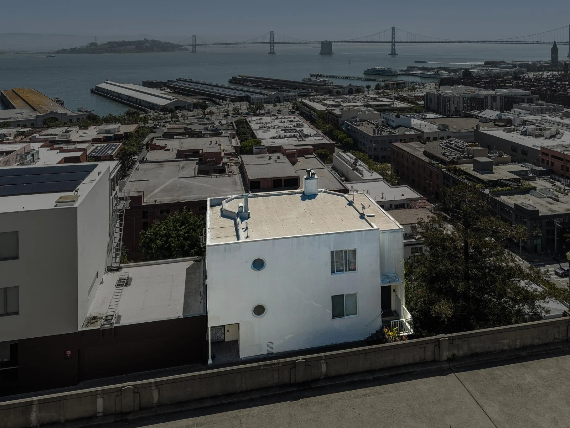 Cityscape with white residential buildings, some with rooftop features, and a view of the bay with docks and boats. A bridge extends across the water in the background under a cloudy sky.