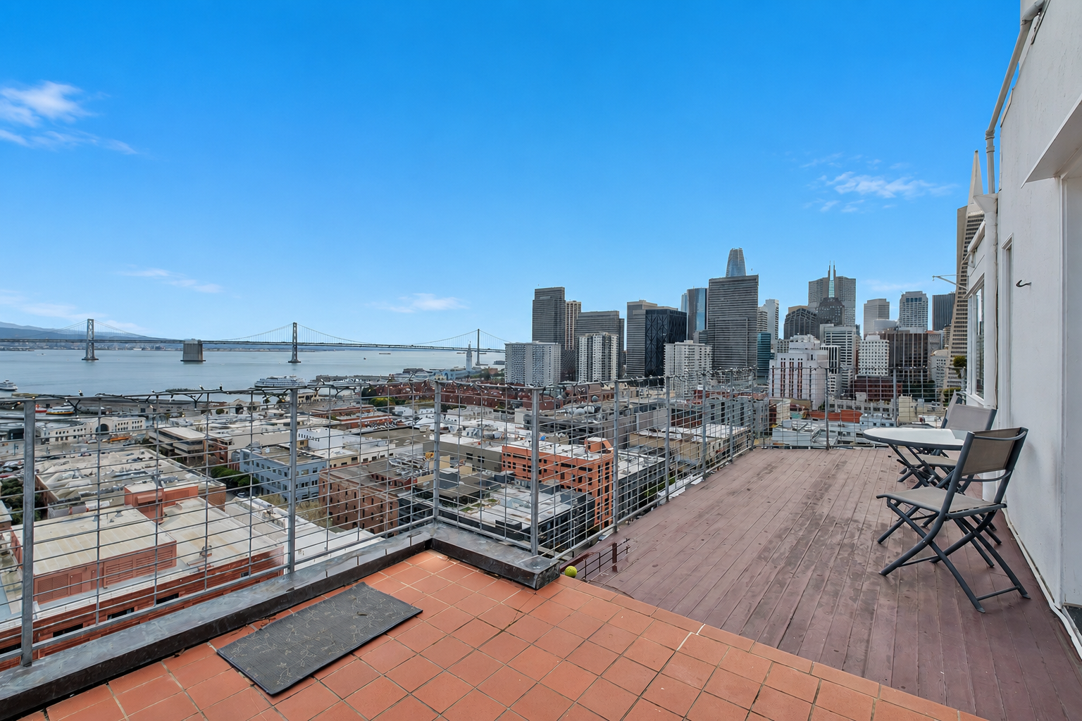 City skyline view from a rooftop balcony with chairs and a table, overlooking the bay and a bridge in San Francisco.