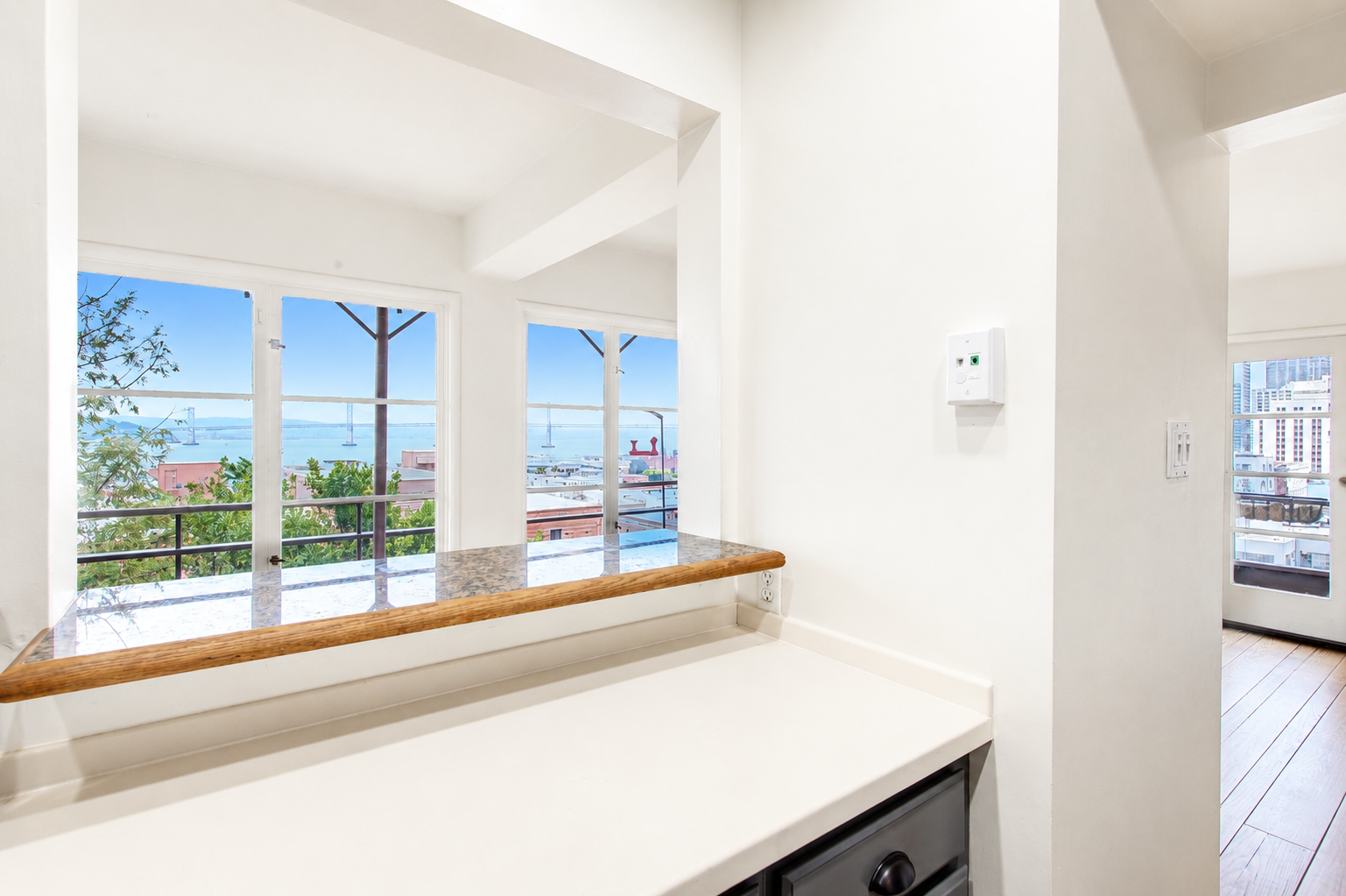 Interior view of a bright room with large windows showcasing a view of a bridge and water. The room has white walls, a granite countertop with a wooden trim, and some electrical outlets.