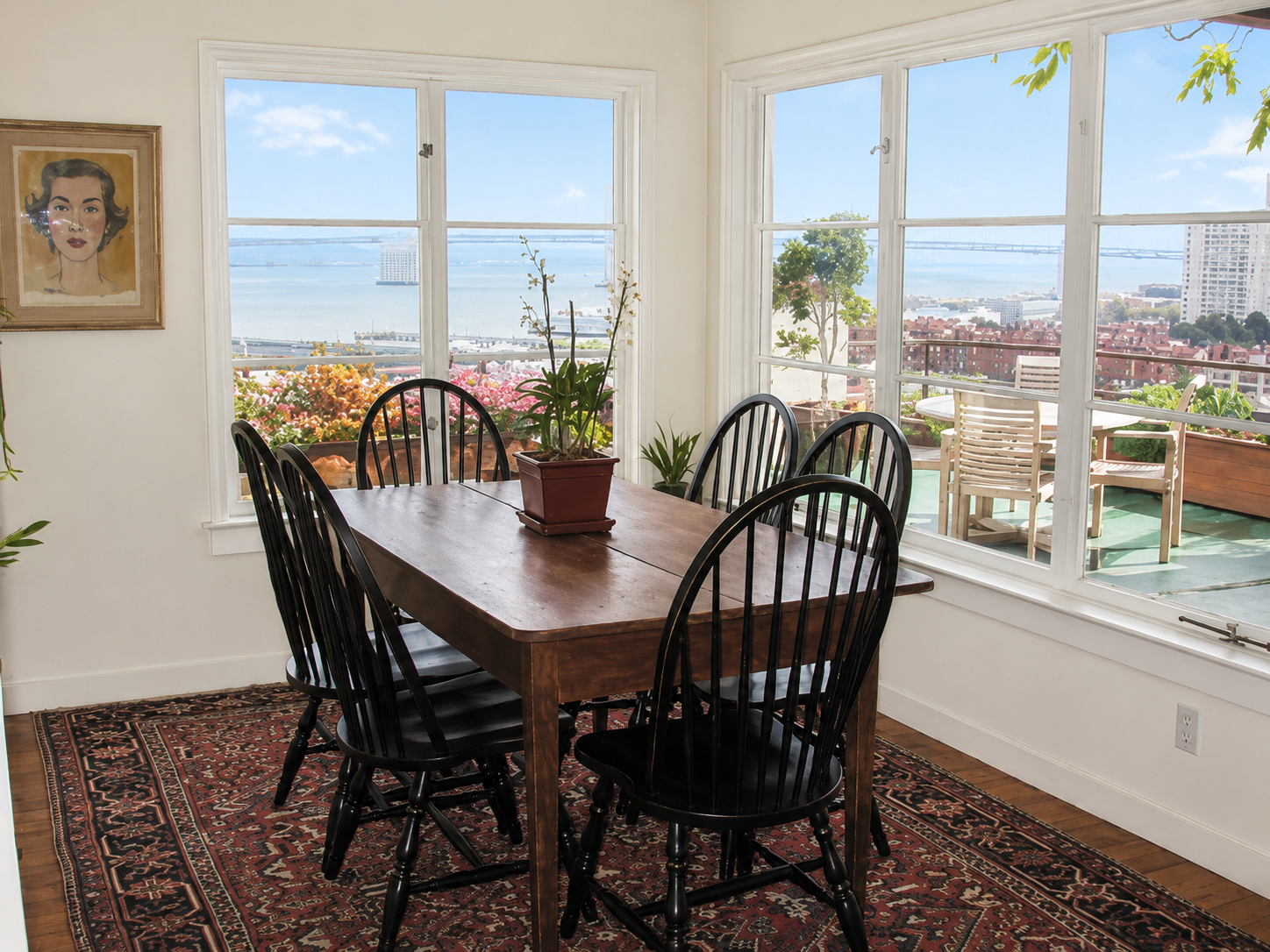 A dining area with a wooden table and six black chairs, located next to large windows with a scenic view of a city, water, and bridges. There are potted plants on the table and outside on the balcony, with blooming flowers and city buildings in the b