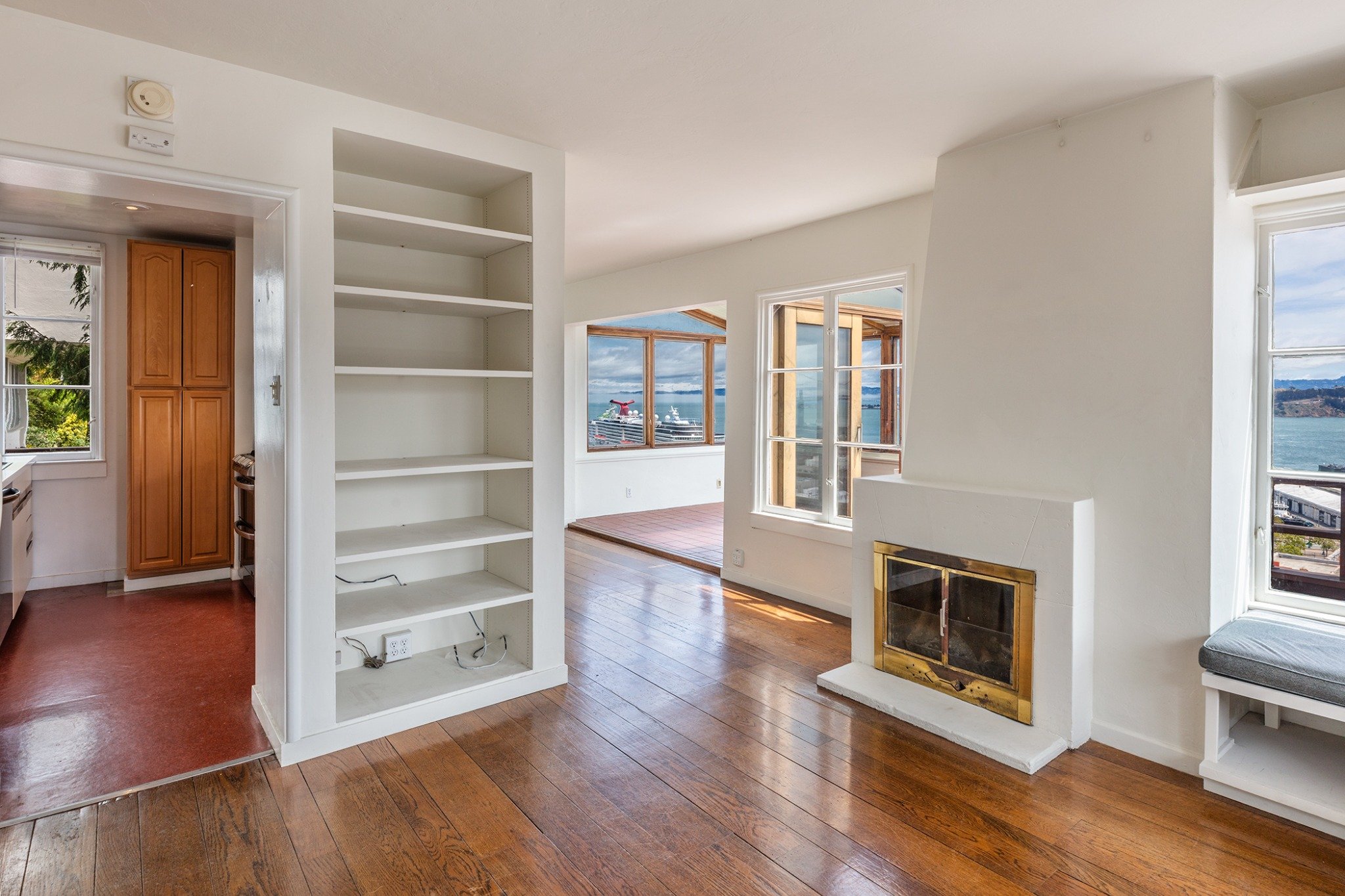 Interior view of a living space with wooden floors, white walls, large windows showing a waterfront view, a fireplace with a golden frame, a built-in white bookshelf, and a doorway leading to a balcony or porch.