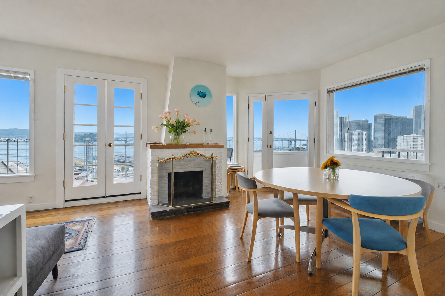 Bright living room with large windows and city view, featuring a white fireplace, a dining table with chairs, and decorative flowers.
