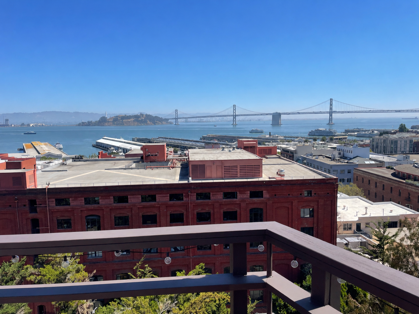 Cityscape view from a balcony overlooking buildings, water, and the San Francisco Bay Bridge on a clear day.