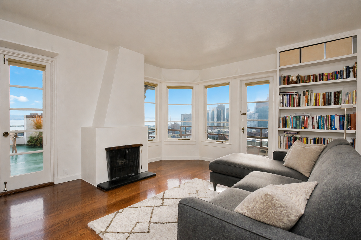Living room with large bay windows showing city skyline, gray sofa with pillows, white bookshelf filled with books, wooden floor, and a white rug.