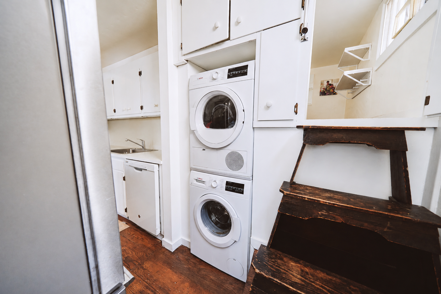 Stacked white washer and dryer units in a small laundry area with wooden stairs next to them. Small kitchen sink with cabinets above and a dishwasher below in the background.