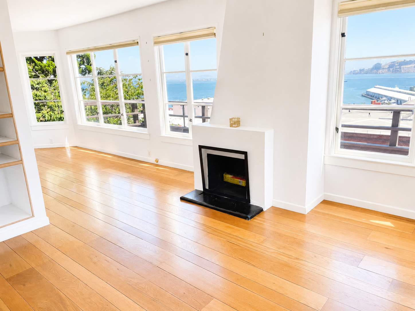 Empty living room with large windows offering ocean view, hardwood floors, white walls, and a fireplace with a decorative box on top.
