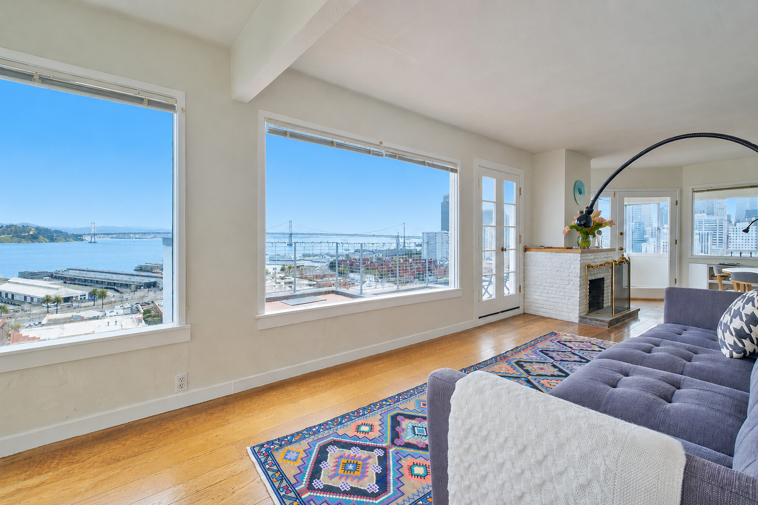 Living room with large windows showing a cityscape and bay, hardwood floors, a patterned rug, purple sofa, and a white brick fireplace.