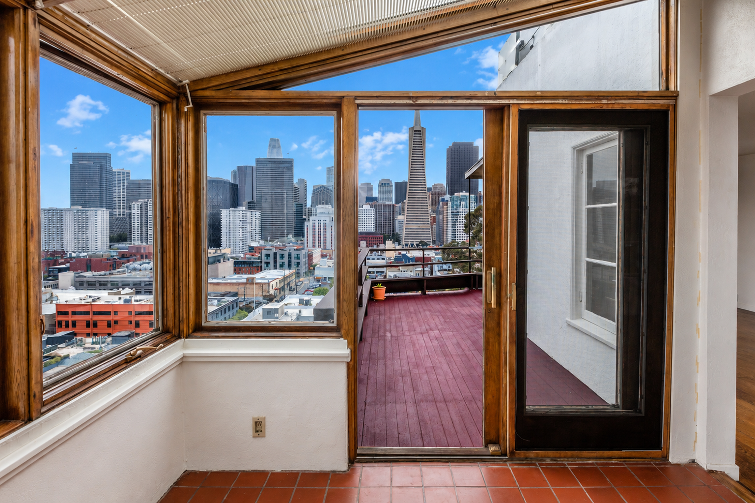 View from inside a room looking out to a city skyline with tall buildings and a prominent triangular-shaped building through large windows and an open balcony with red wooden flooring.