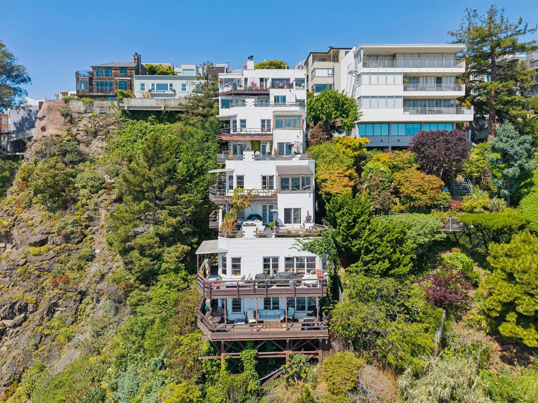 Aerial view of hillside residential buildings and lush greenery next to a rocky cliff on a sunny day.