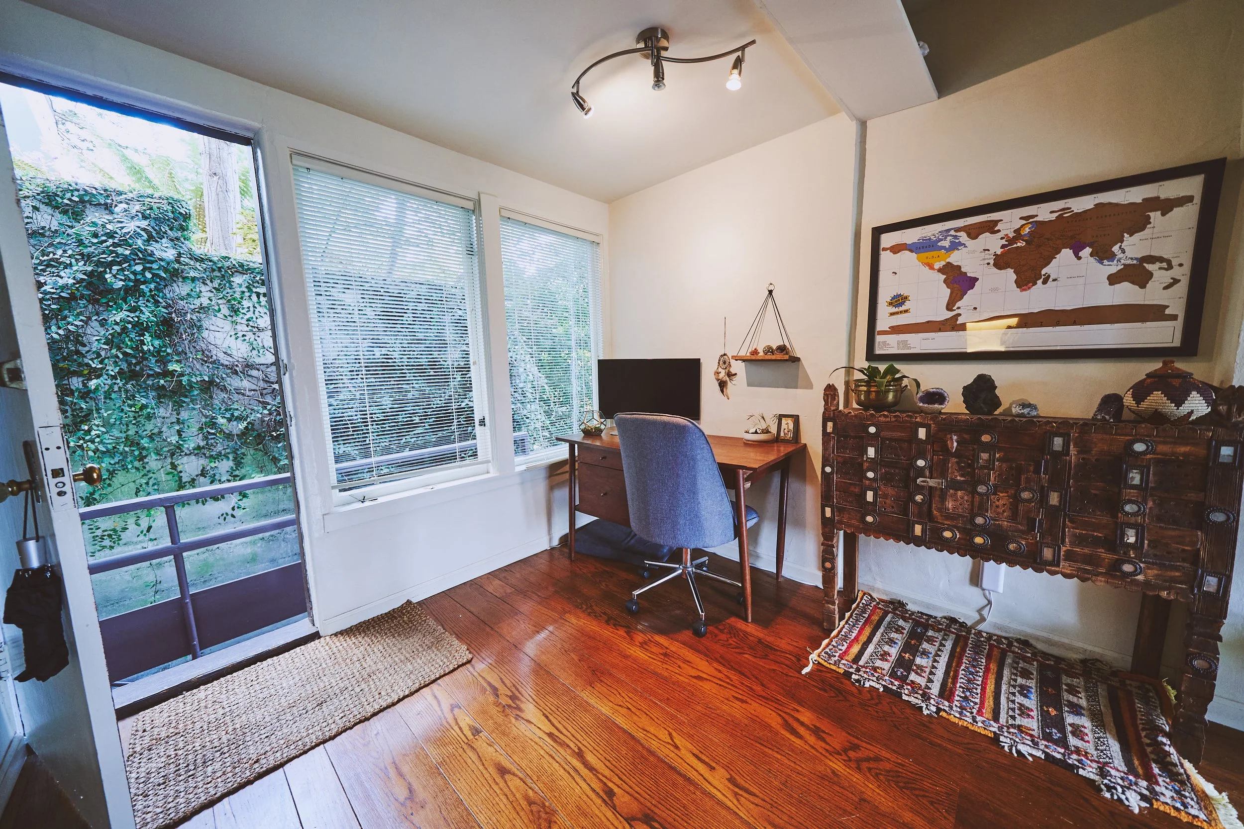 A cozy room with wooden floors, a small wooden desk, a blue office chair, a large world map on the wall, a wooden cabinet with decorative items, and large windows with blinds that overlook a garden.