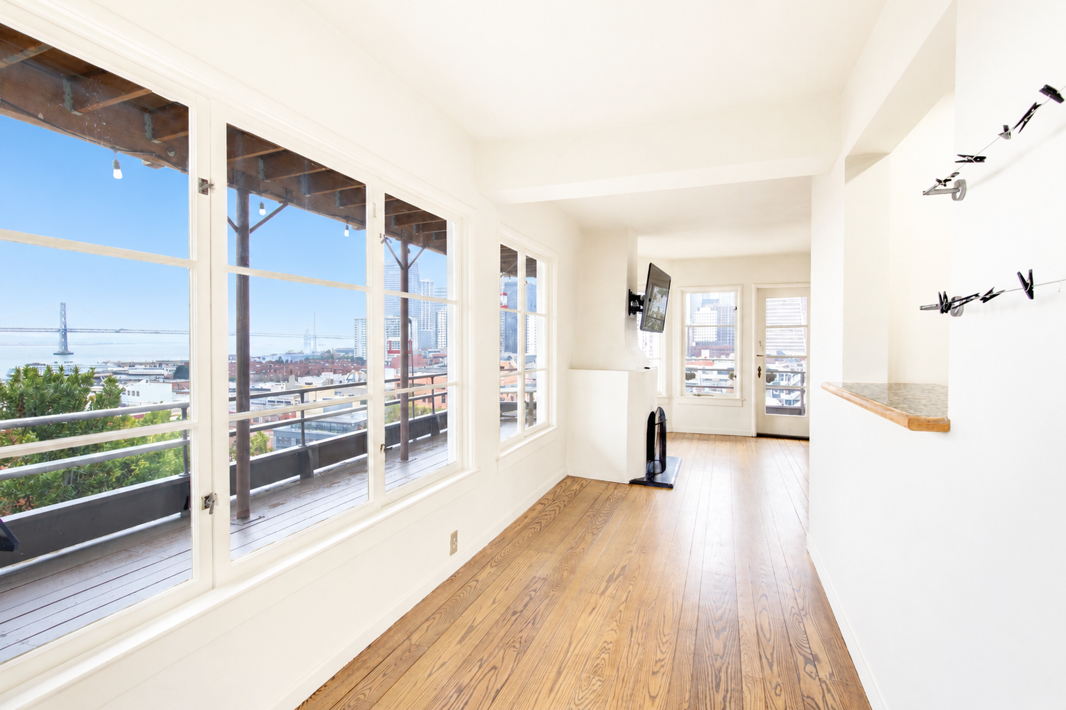 Bright living room with large windows showing cityscape and bridge view, hardwood floors, wall-mounted TV, small fireplace, and a door leading to a balcony.