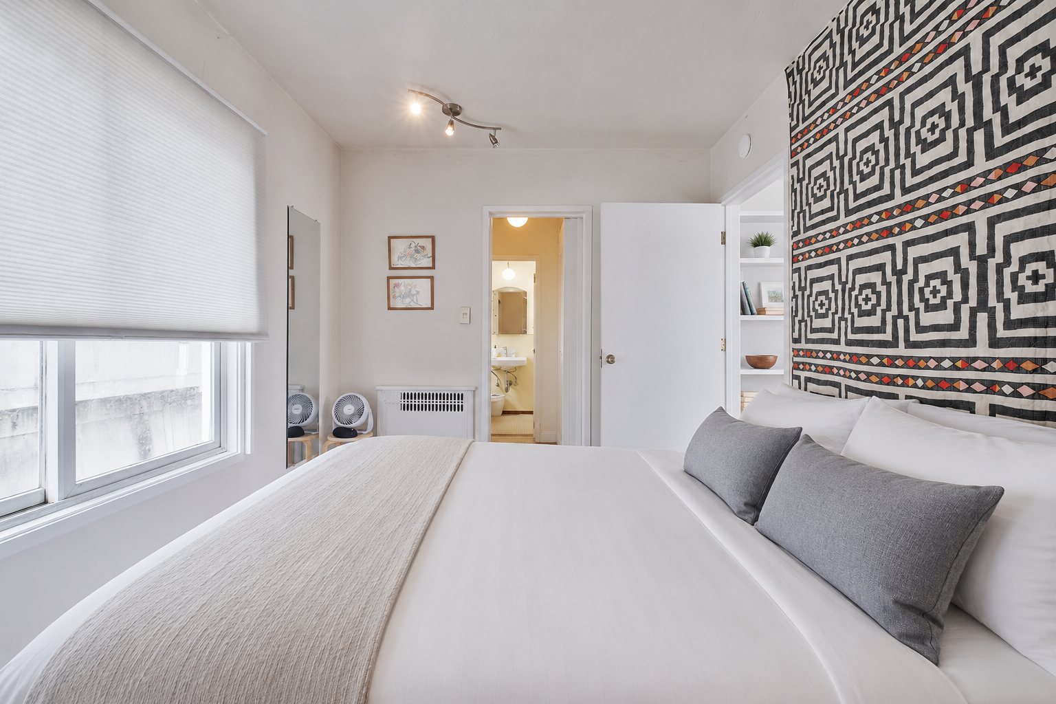 A bedroom with a large bed, gray and white pillows, a patterned wall hanging, and a window with a closed white blind. There is a door leading to a bathroom, and small fans on the floor near the wall. The room has white walls and built-in shelves with