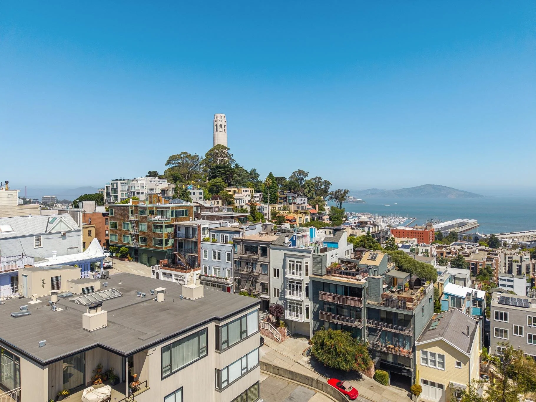 Aerial view of a hilly hillside neighborhood with modern multi-story houses, some with balconies and rooftop decks, nestled among trees. The Bell Tower, a tall, white, historic structure, stands prominently atop the hill. Beyond the neighborhood, the