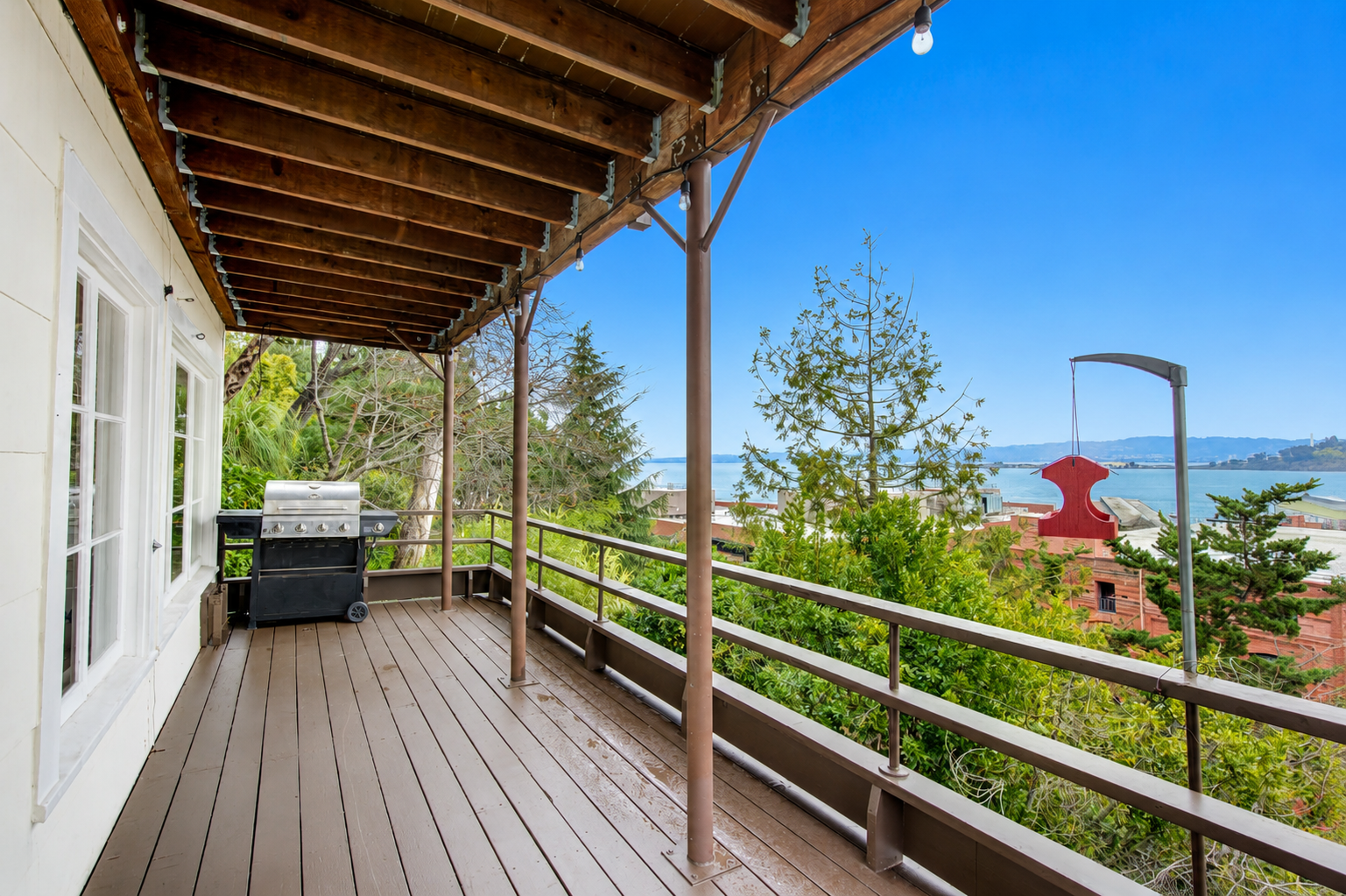 A balcony with a wooden floor and an overhead wooden ceiling, featuring a barbecue grill and string lights, overlooking a view of trees, rooftops, water, and hills in the distance under a clear blue sky.