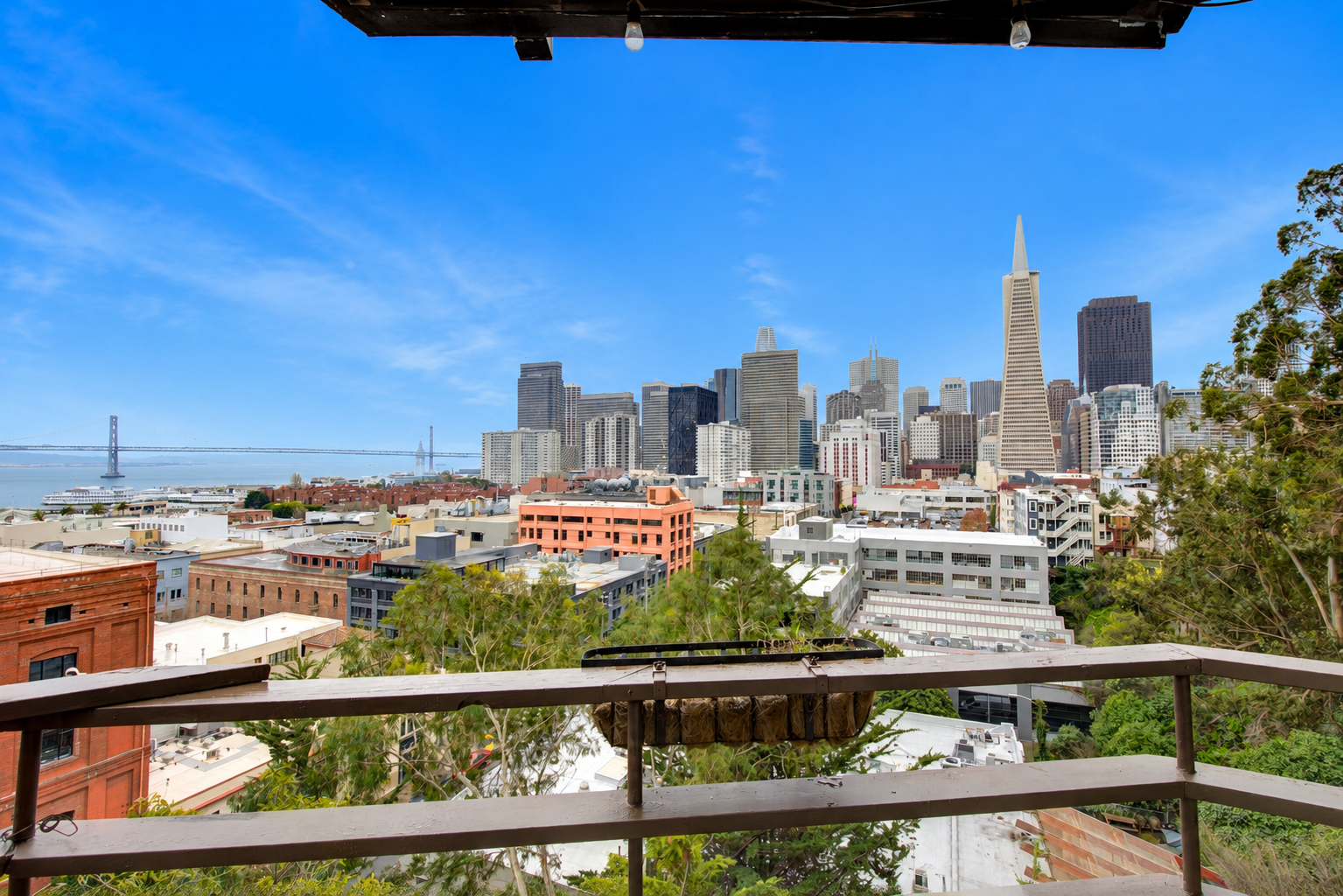 View of the San Francisco skyline with a clear blue sky, including the Transamerica Pyramid, from a balcony with a railing and some greenery in the foreground.