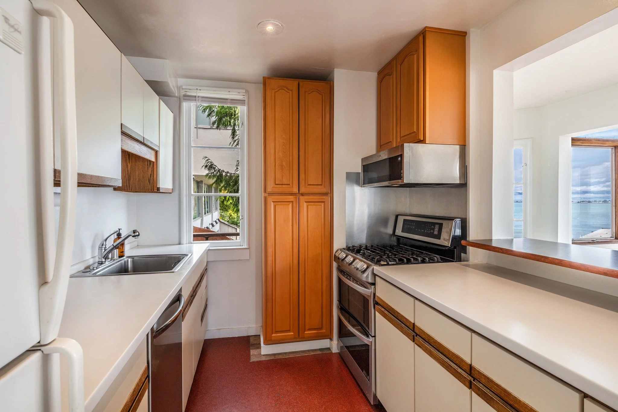 A small kitchen with white countertops, wooden cabinets, a stainless steel microwave, and a gas stove. There is a window showing trees outside and a sliding glass door leading to a balcony.