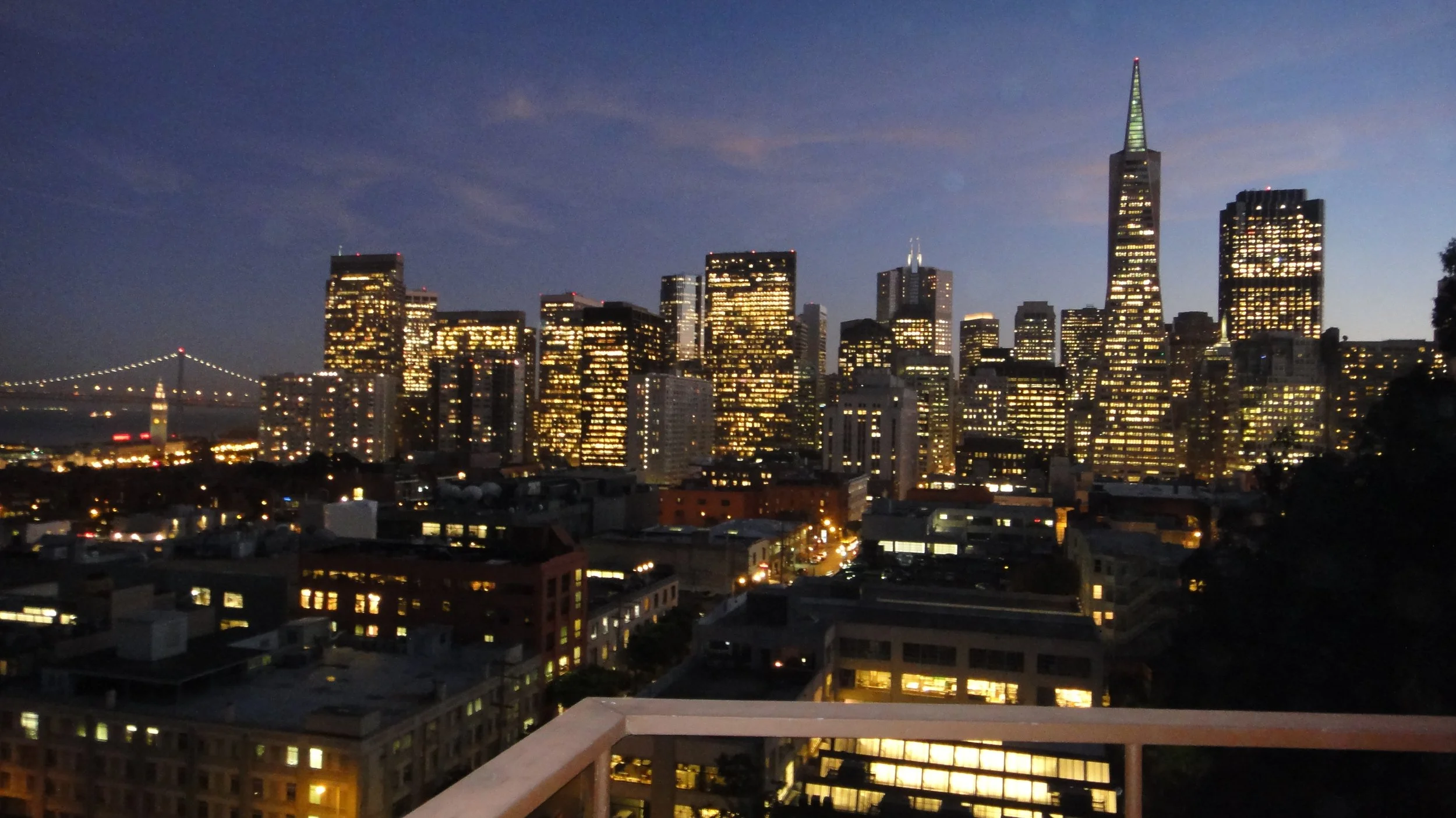 Nighttime city skyline with illuminated buildings, including a tall skyscraper with a pointed top, and a bridge with lights over water in the background.