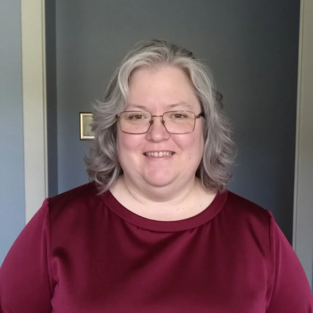 A woman with glasses and gray hair smiling at the camera, wearing a maroon top, in a room with dark blue walls.