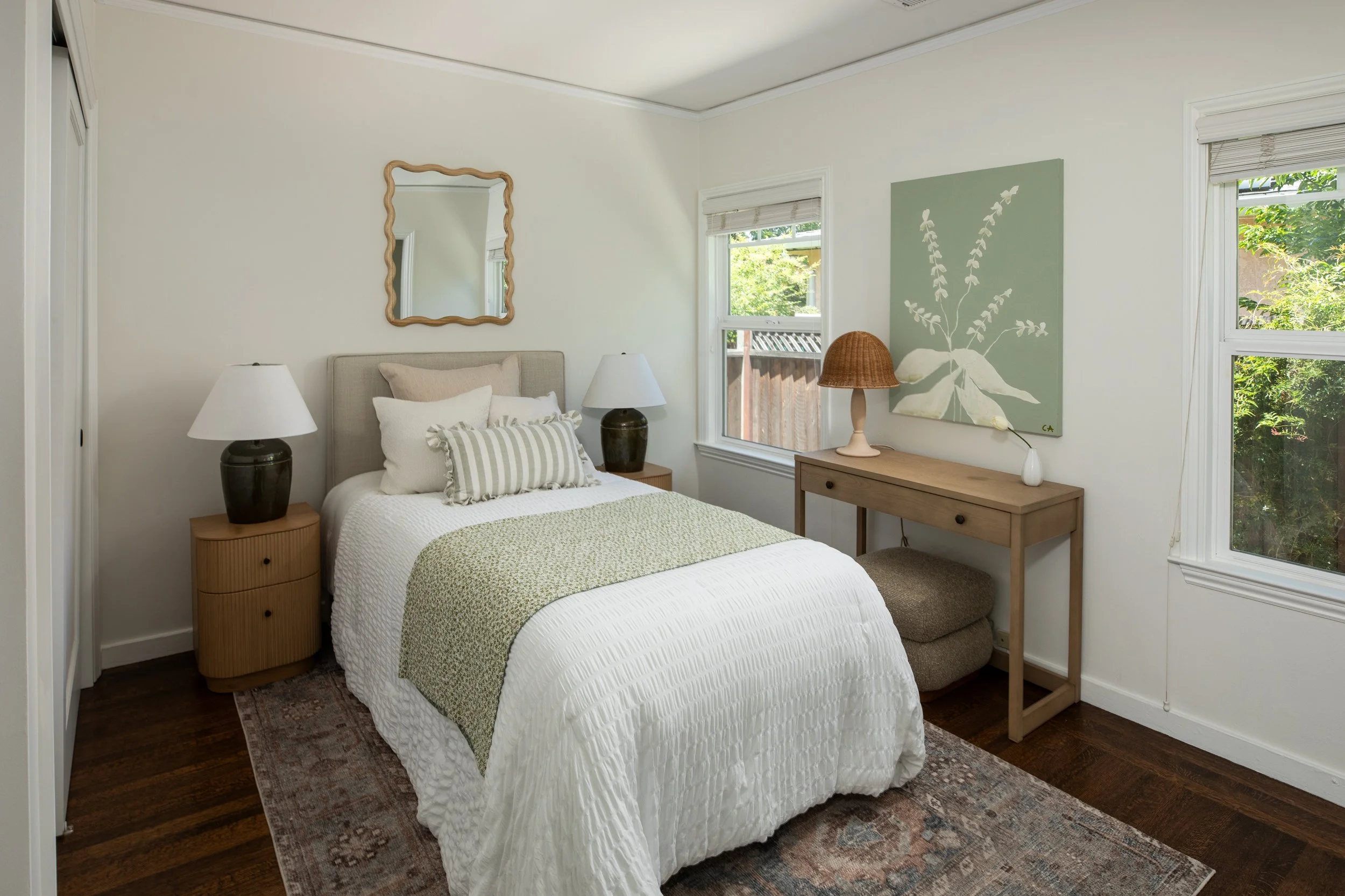 A cozy bedroom with a bed, matching striped pillows, and a green patterned bedspread. On the left, there's a wooden nightstand with a large black lamp, and on the right, a wooden dresser with a small white lamp featuring a woven shade. The walls are 