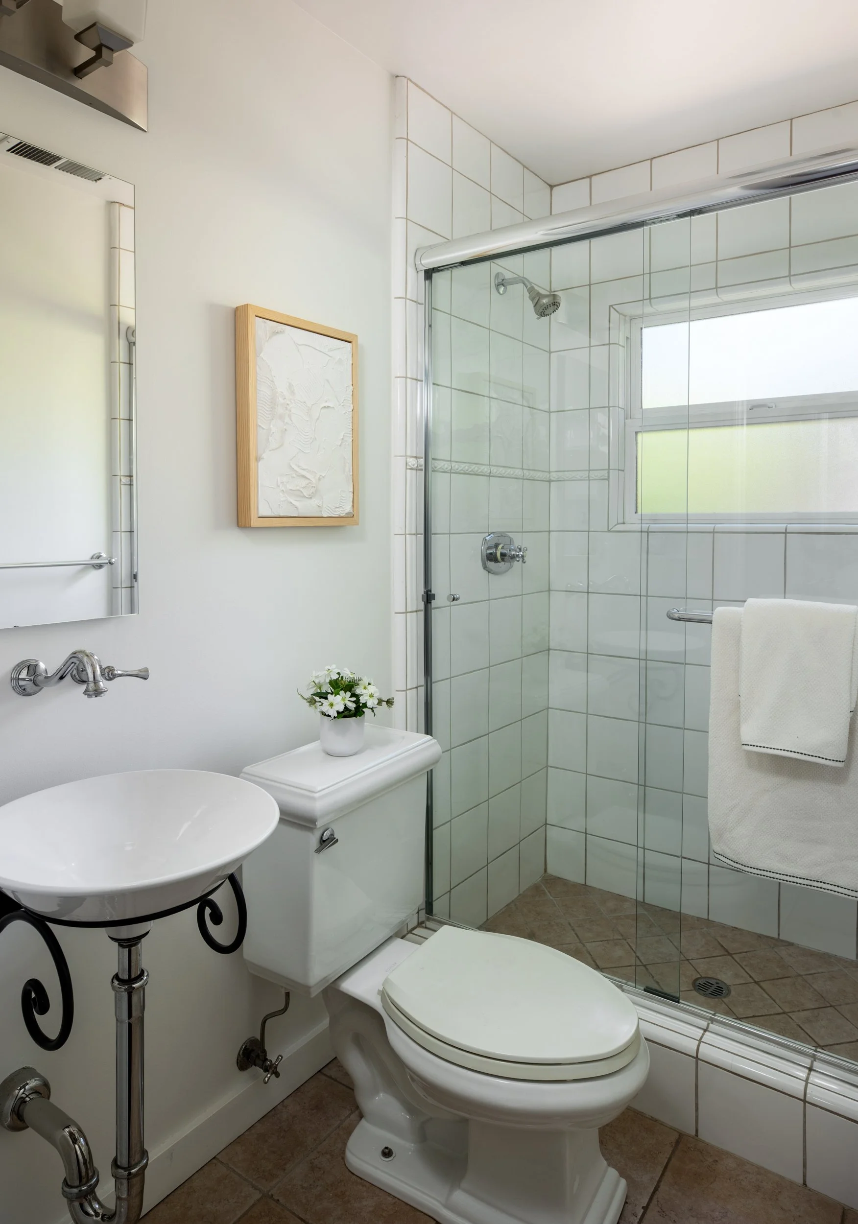 Bathroom with white walls, a toilet, a vessel sink on a black wrought iron stand, a wall mirror, a small white side table with a flowerpot, a framed white textured artwork, a towel rack, and a glass shower enclosure with a window.
