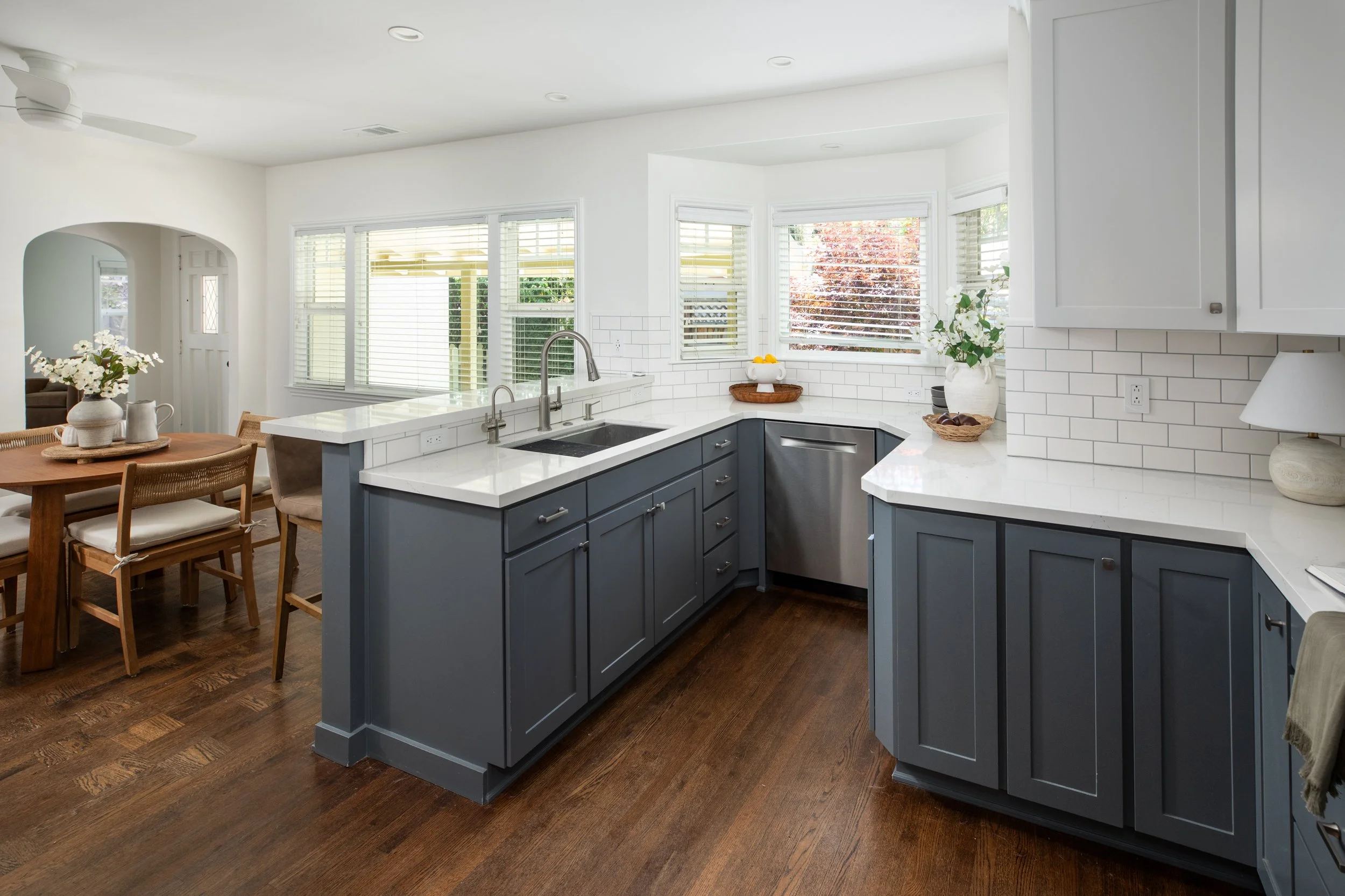 Modern kitchen with white countertops, blue cabinets, white subway tile backsplash, and large windows with blinds, with a view into a dining area with a wooden table and chairs.