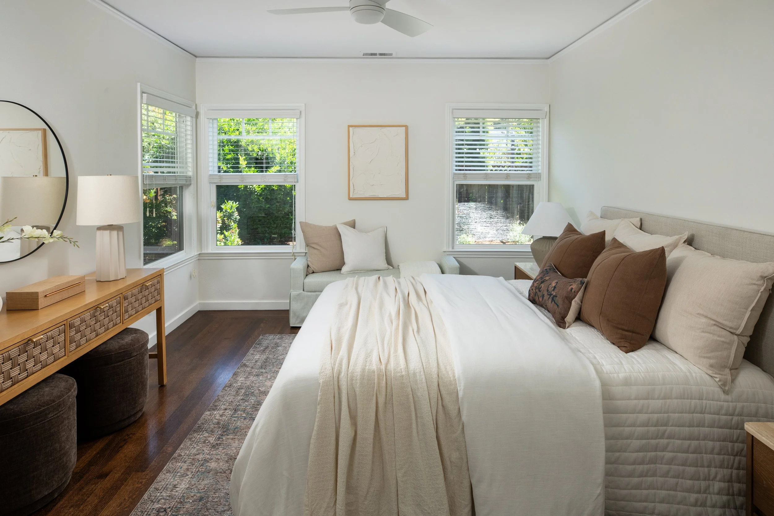 Bright bedroom with white walls and three large windows with white blinds, a bed with beige and brown pillows, a bedside lamp, a wooden dresser, and artwork on the wall.