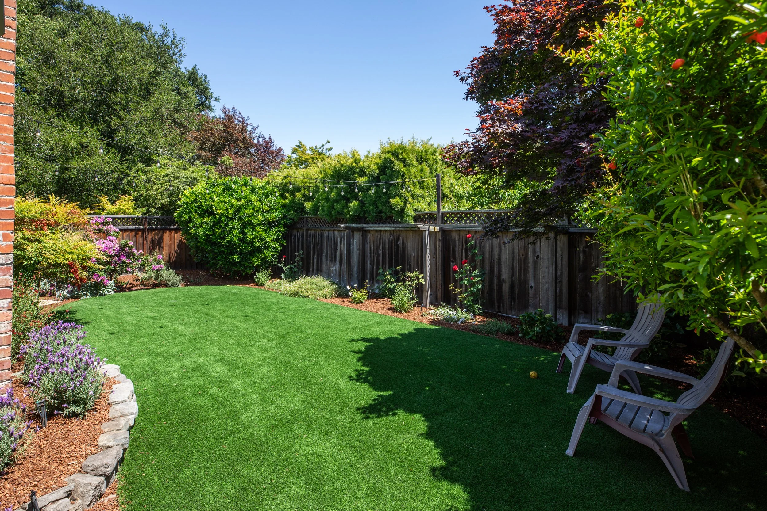 A backyard garden with vibrant green grass, colorful flowering plants, and a wooden fence. There are two white rocking chairs on the right side, with a garden on the left and various trees and bushes in the background under a bright blue sky.