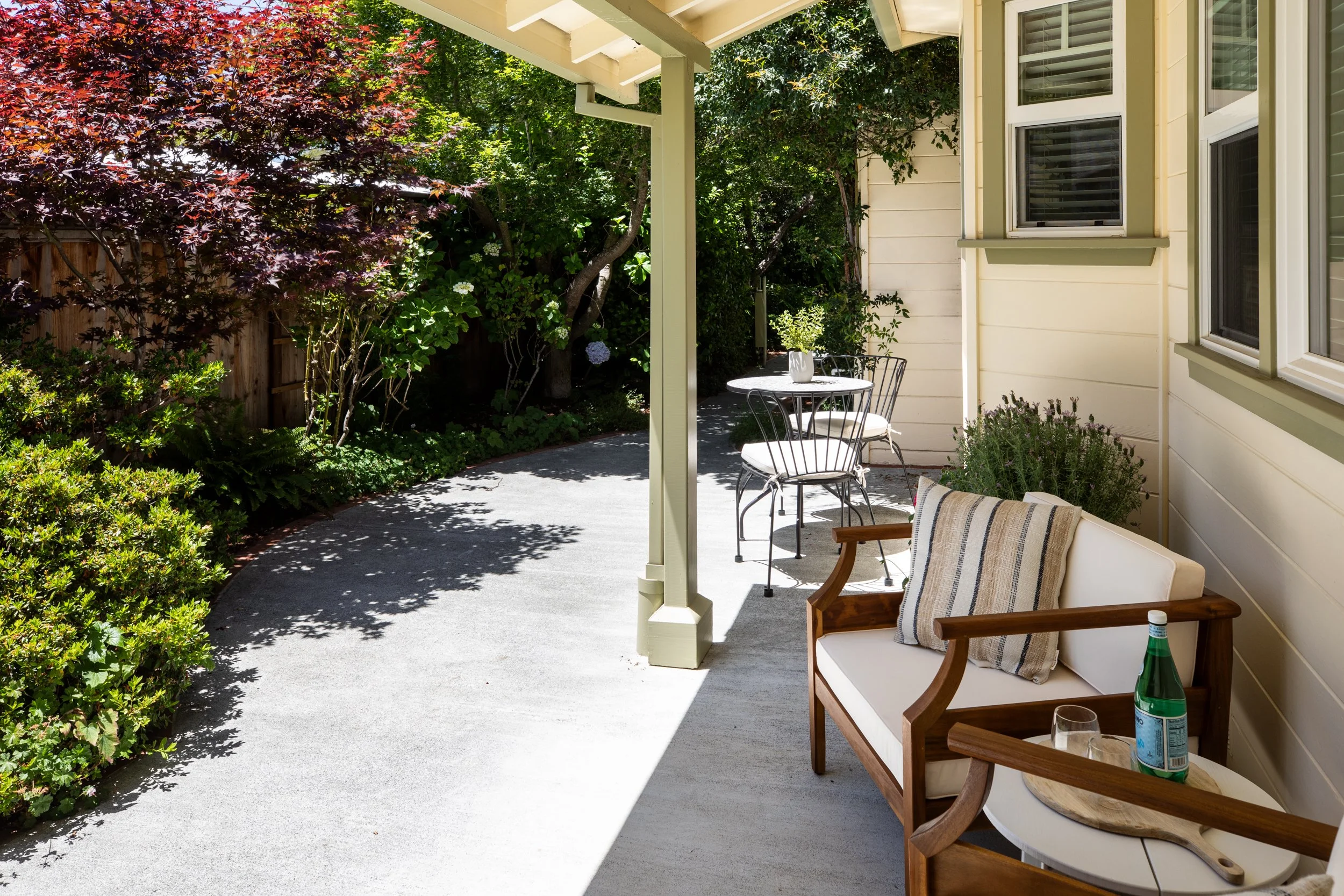 A cozy covered porch with outdoor seating, including a wooden bench with striped cushions, a small round table with two metal chairs, and greenery surrounding the area, including trees and bushes, during daytime.