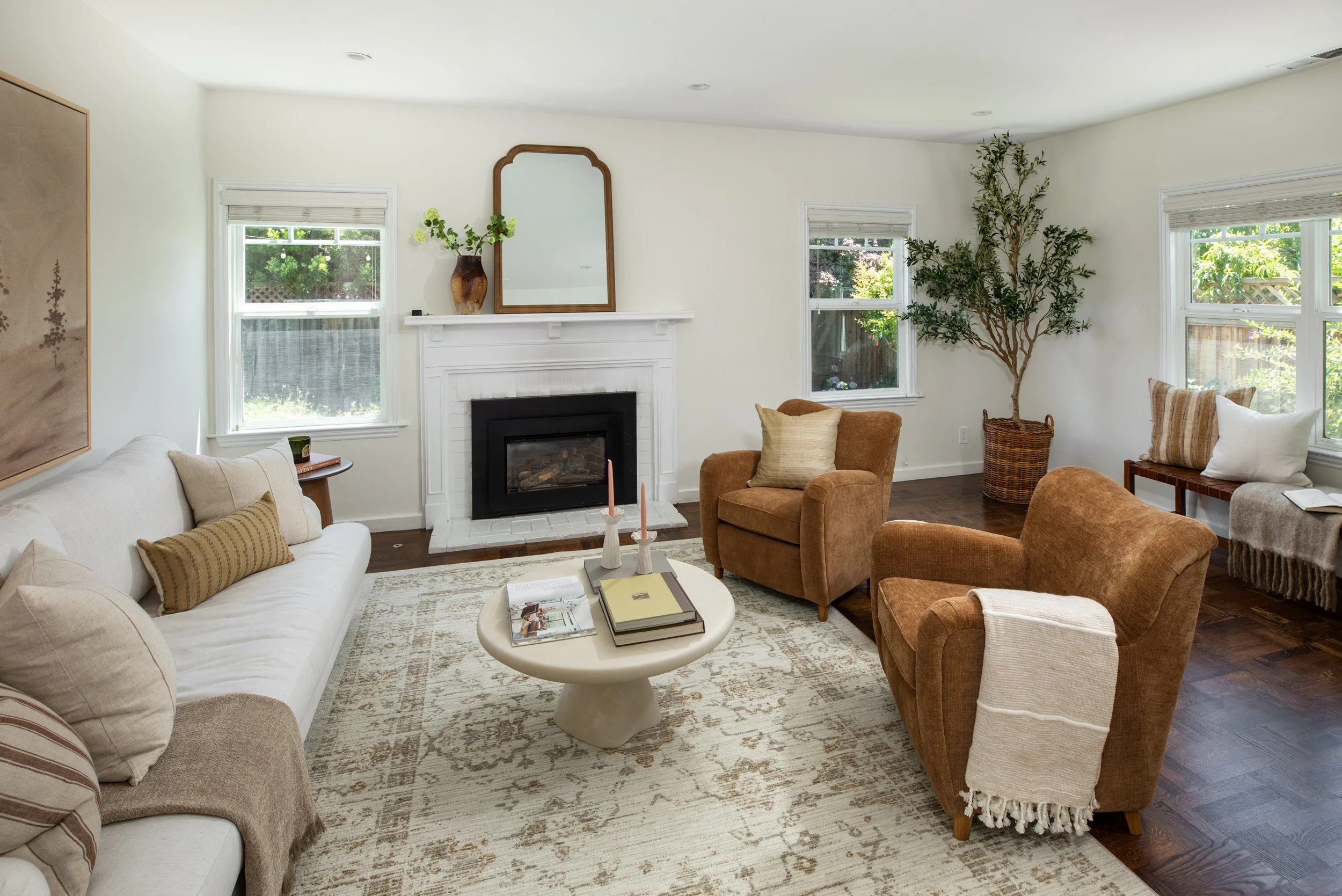A cozy living room with cream-colored sofa and armchairs, a white coffee table with books and candles, a fireplace with a mirror and flower vase above it, large windows with blinds, a tall potted plant, and a patterned area rug.