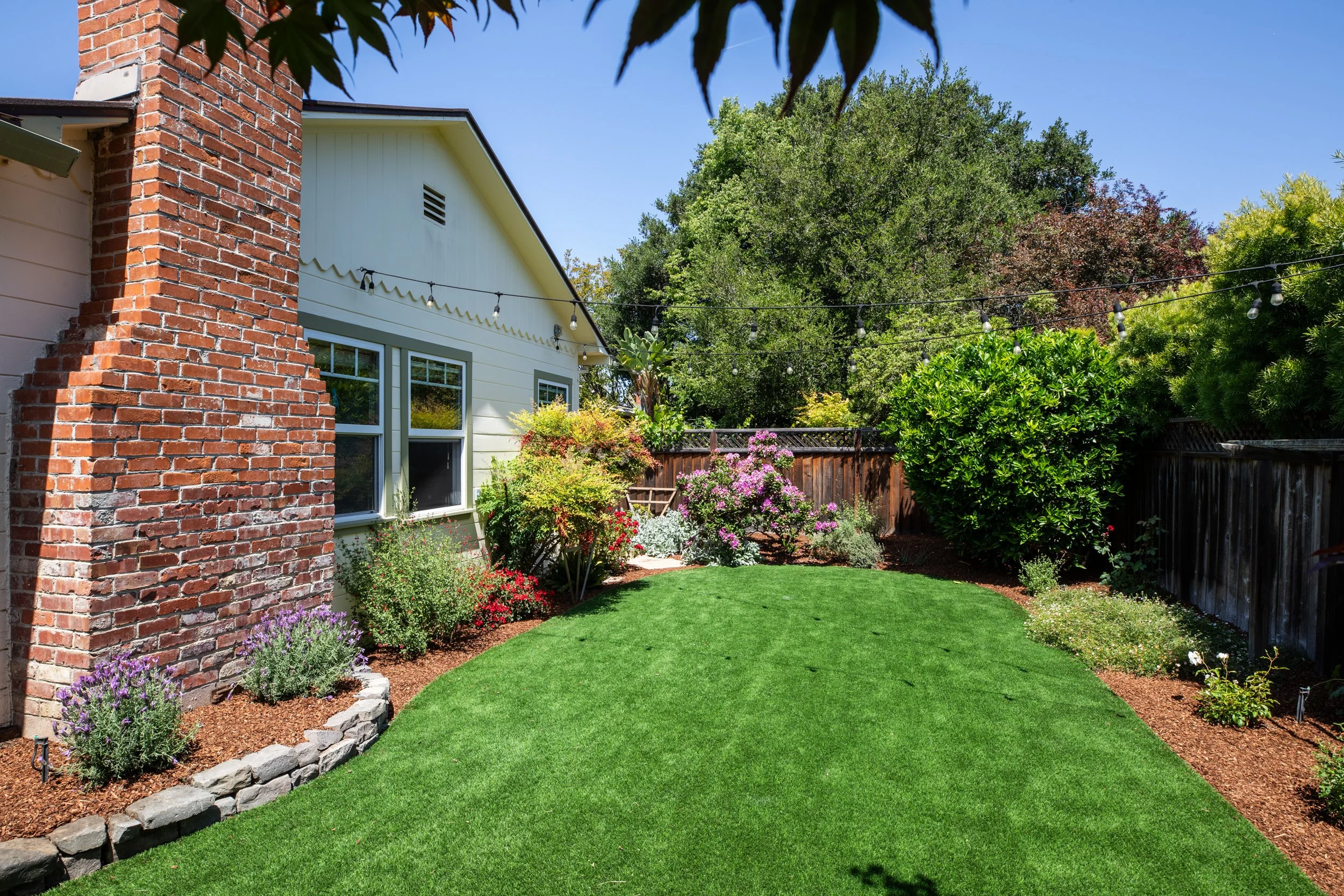 A well-maintained backyard garden with a lush green lawn, colorful flowers, and bushes outlined with stone edging, with a white house and trees in the background, under a clear blue sky.