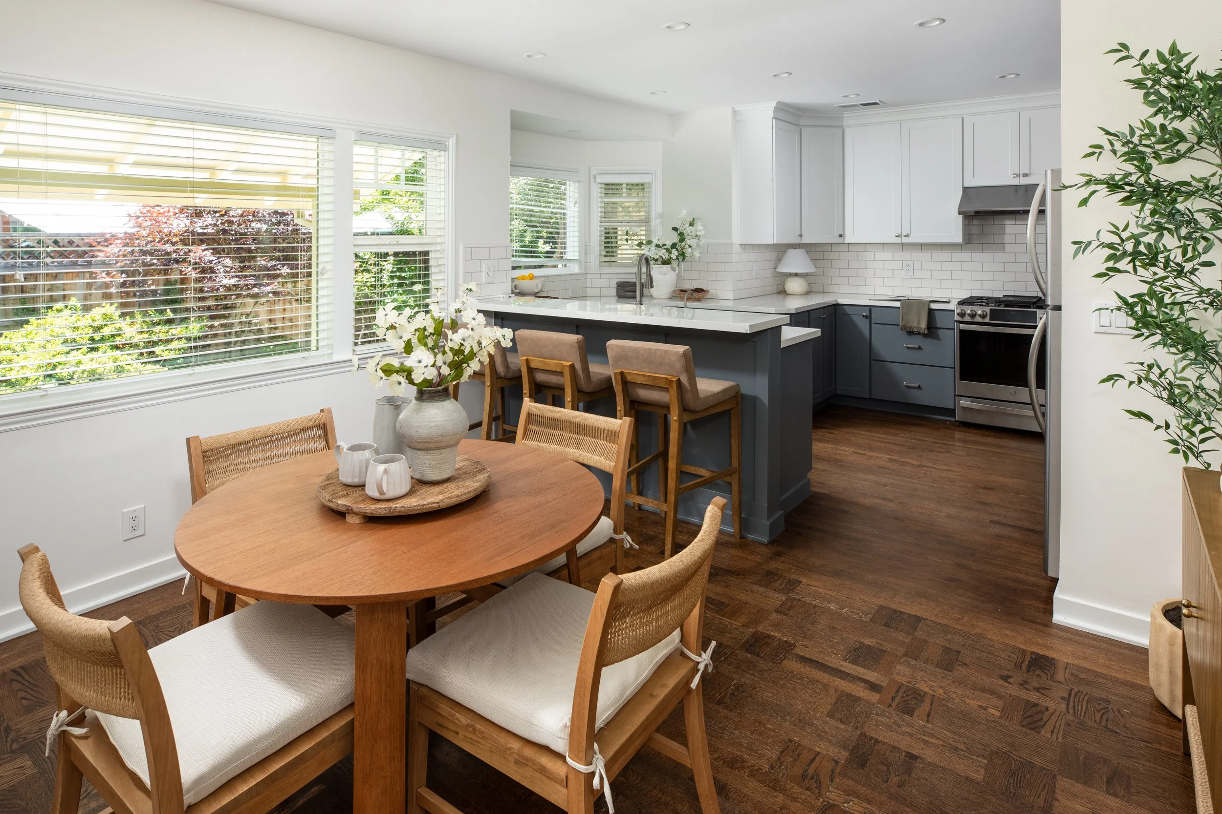 A bright kitchen with large windows, white cabinets, a white subway tile backsplash, stainless steel appliances, a kitchen island with bar stools, and a round dining table with a flower centerpiece and wooden chairs.