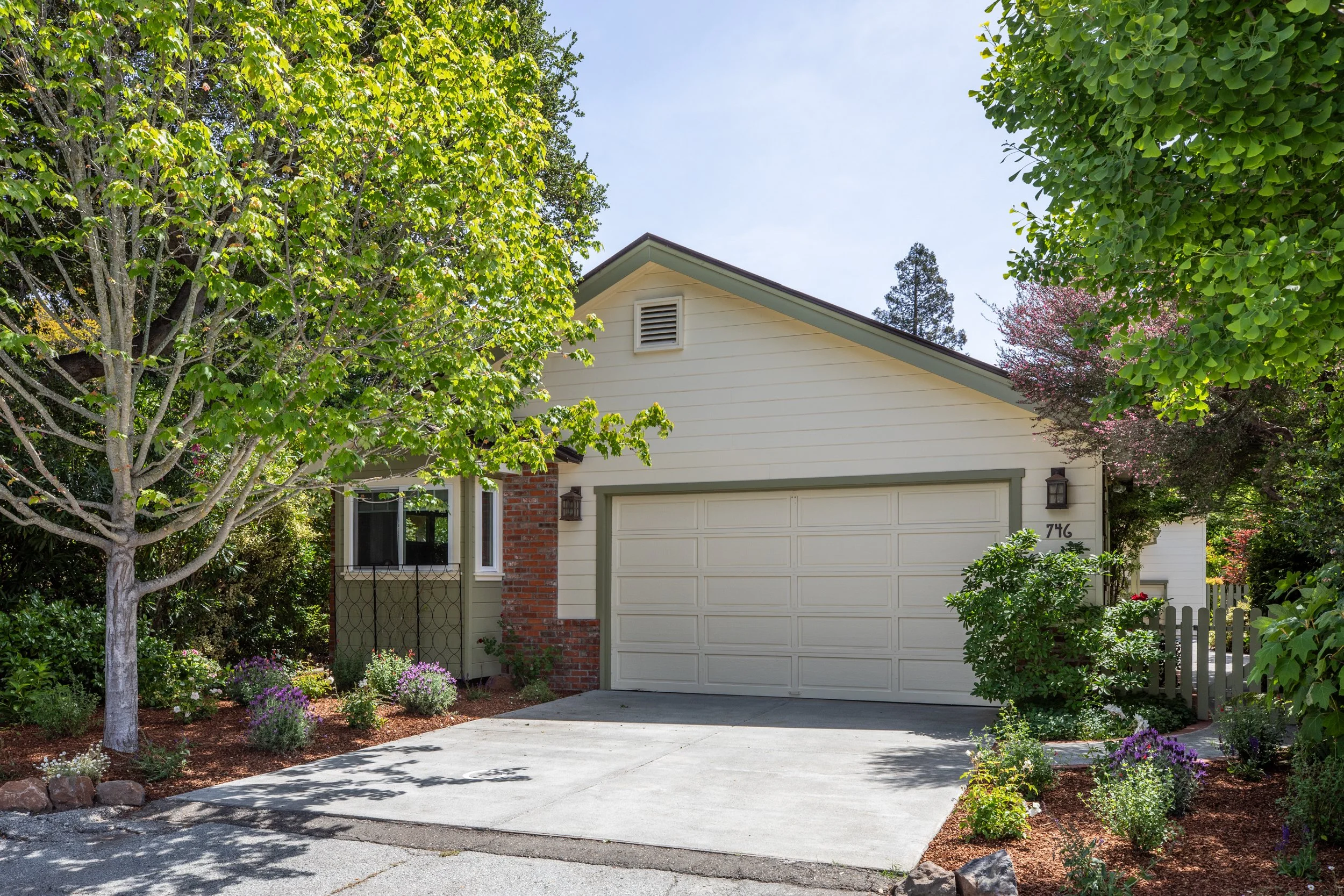 A suburban house with a white garage door, surrounded by greenery including a large leafy tree on the left and flowering trees on the right, and a neat front yard with plants and flowers.