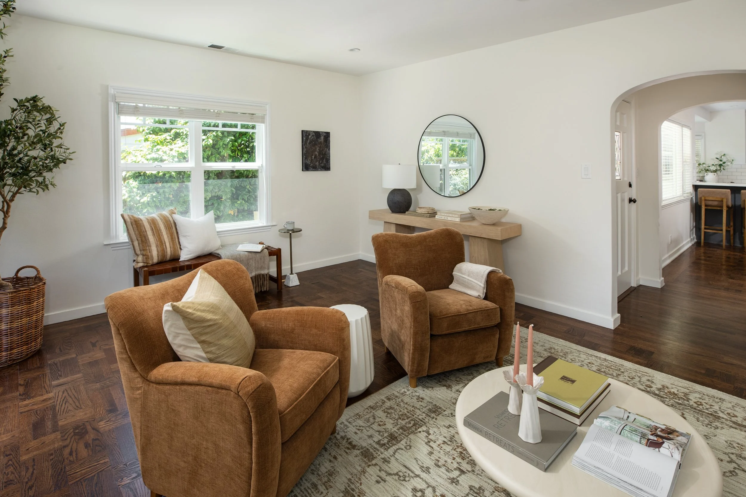 A living room with beige upholstered chairs, a white coffee table with books and candles, a wire basket, a large window with greenery outside, and decorative items like a mirror, lamp, and artwork.