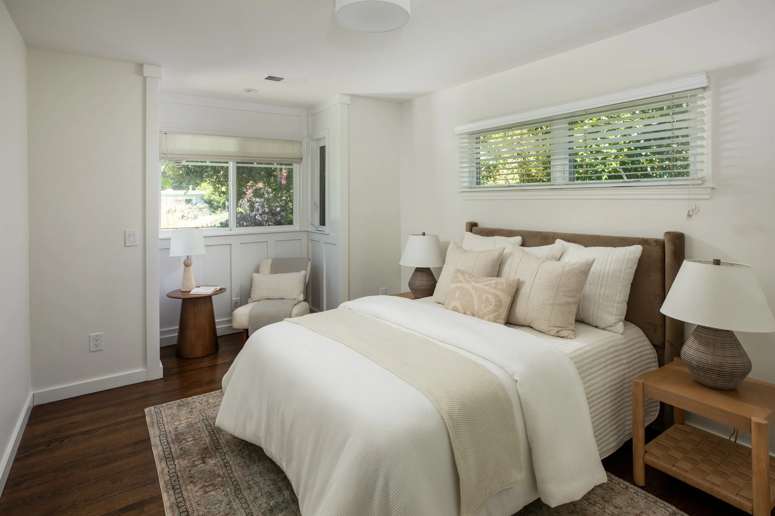 A bedroom with a white bed, beige pillows, a bedside table, and a window with blinds showing greenery outside.