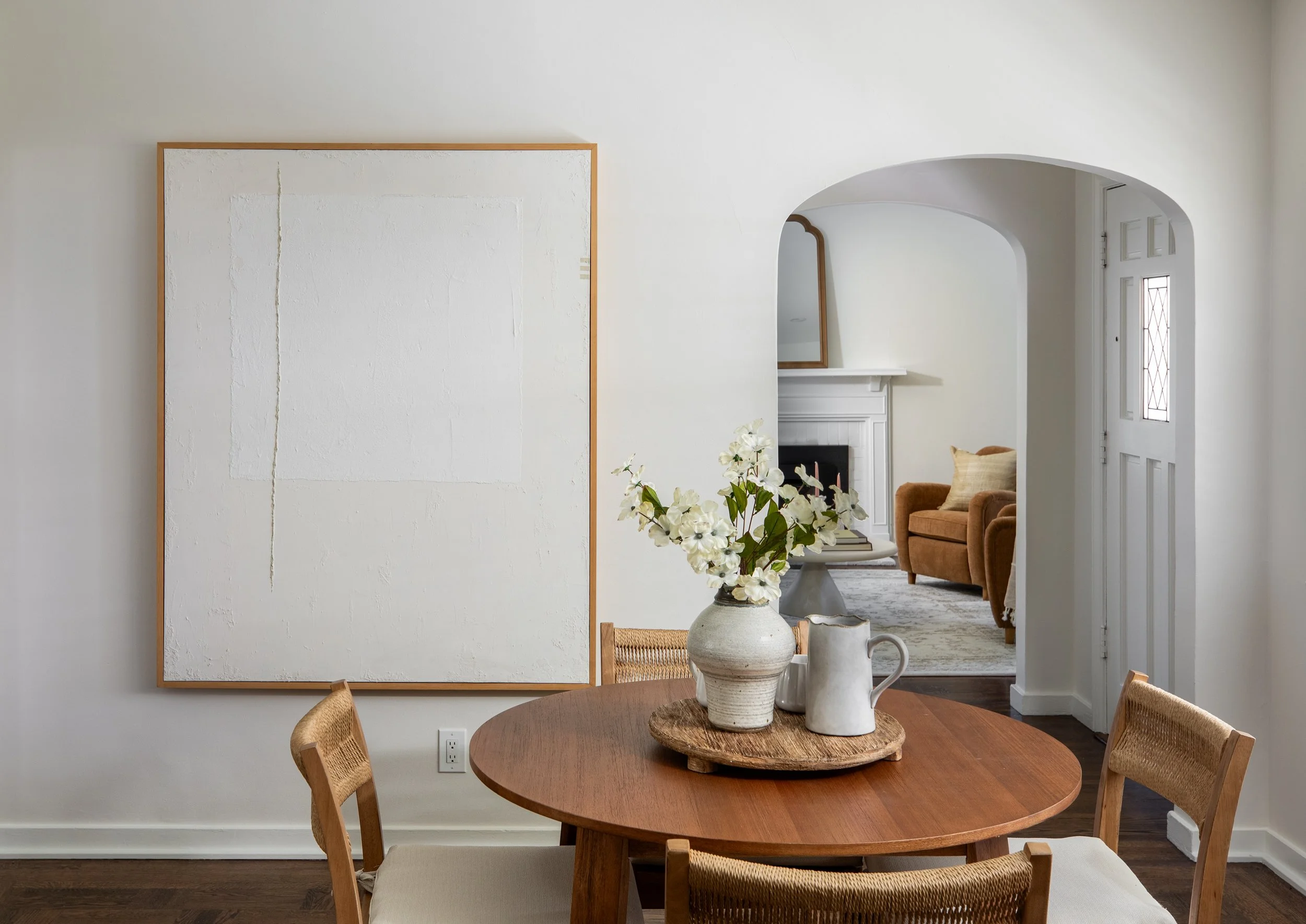 A dining room with a wooden round table and four wicker chairs. On the table, there's a tray with decorative white vases and a flower arrangement with white flowers. In the background, there's a white fireplace, a mirror, a brown armchair, and a wind