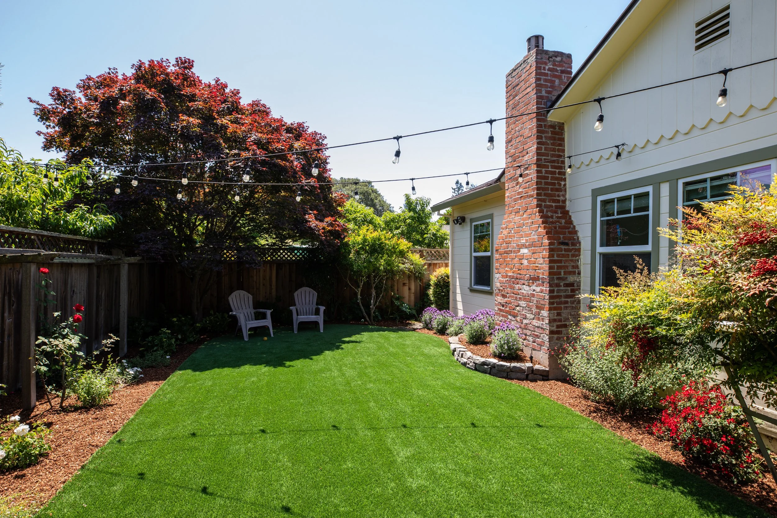 A backyard with a green lawn, flowering bushes, a wooden fence, two white chairs, a tree with red leaves, and string lights hanging above. Part of a house with a brick chimney and window is visible.
