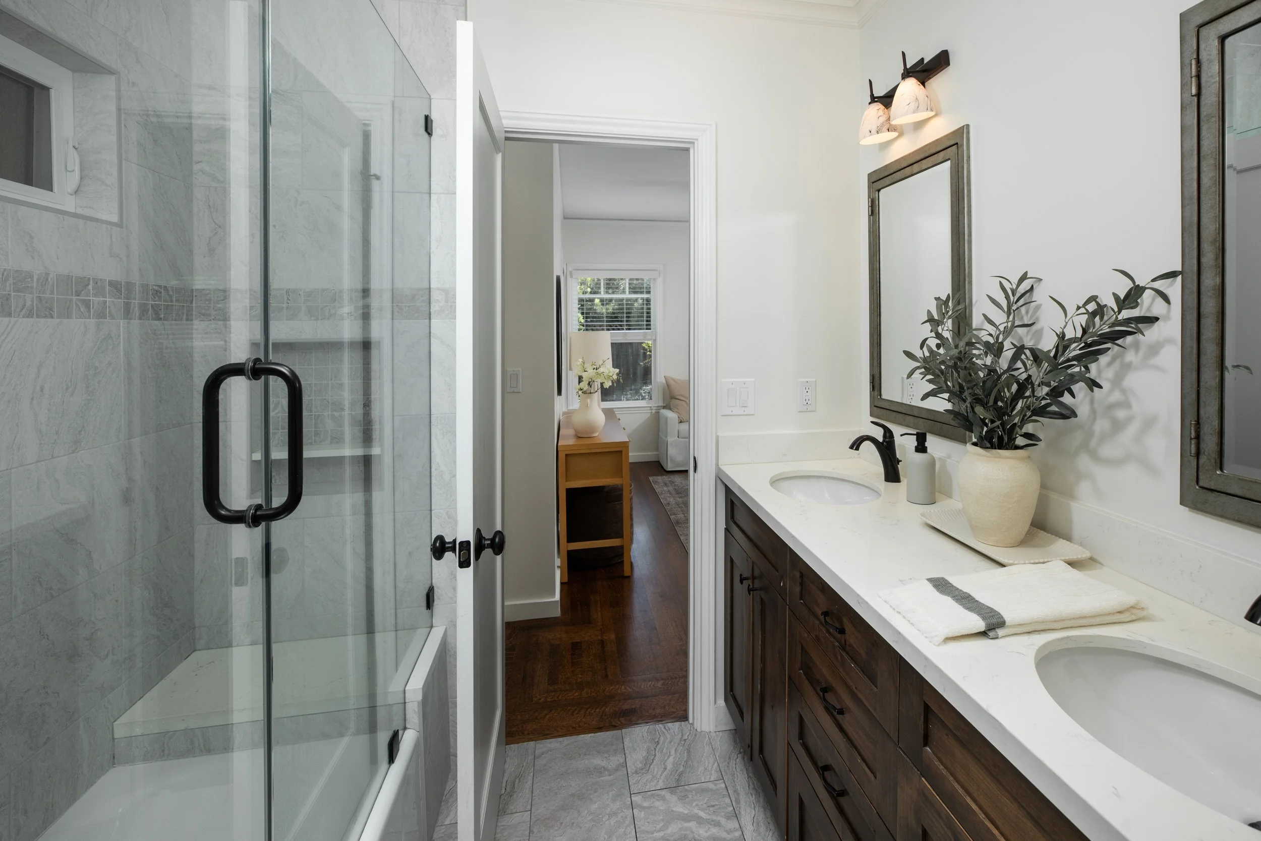 A bathroom with a glass shower, a white vanity with a dark wood cabinet, a large mirror, and a potted plant. Visible doorway leading to a room with windows and a small table.
