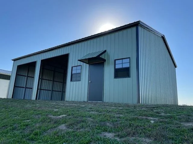 A large metal building with two garage doors and a smaller door between them, situated on a grassy area under a clear sky.