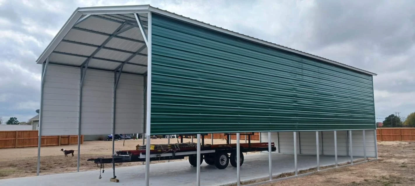 A large metal shed under construction on a concrete pad with a trailer beneath it, surrounded by a wooden fence and a cloudy sky.