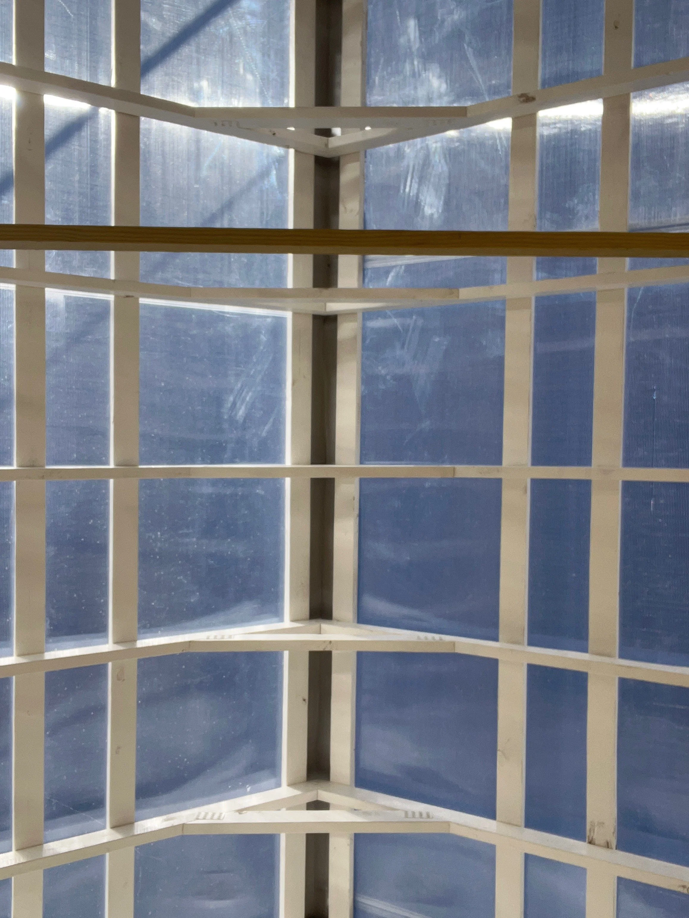 Interior view of a large, multi-pane window with a grid of white wooden frames, showing a daytime outdoor scene with a blue sky and some clouds.