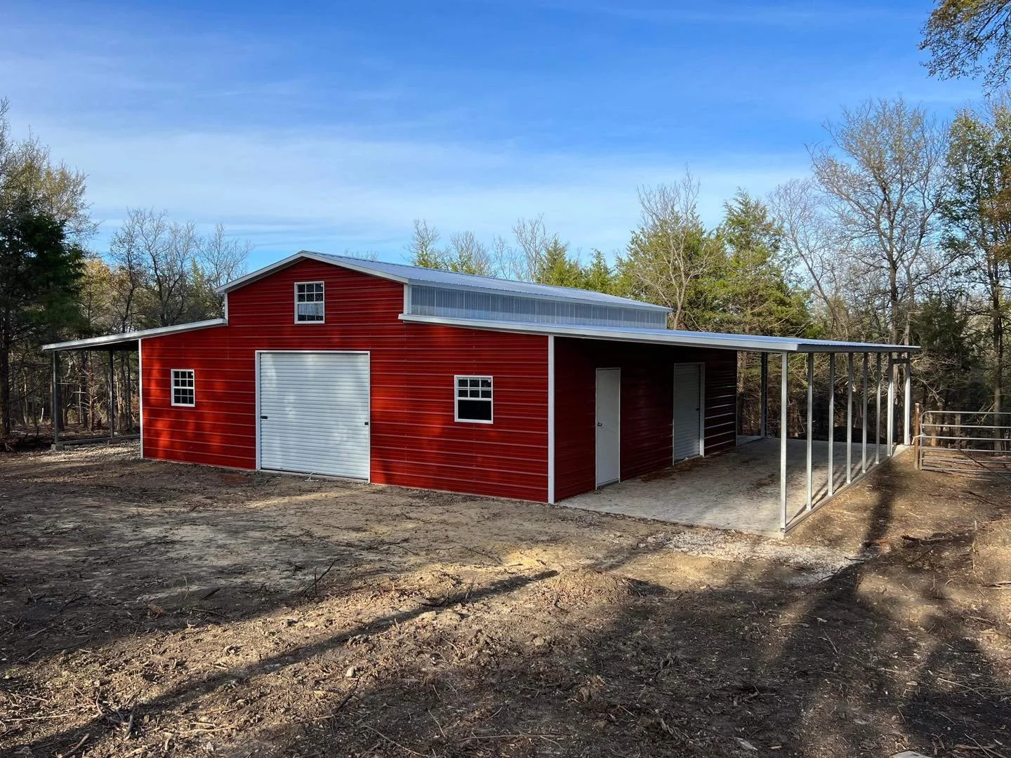 Red barn with a metal roof, small windows, and a large garage door, situated on a dirt lot with trees in the background.
