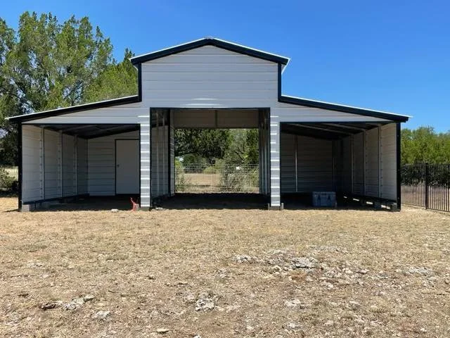 A metal carport with a central open section and two side enclosed sections, situated on a grassy area with a clear blue sky and trees in the background.