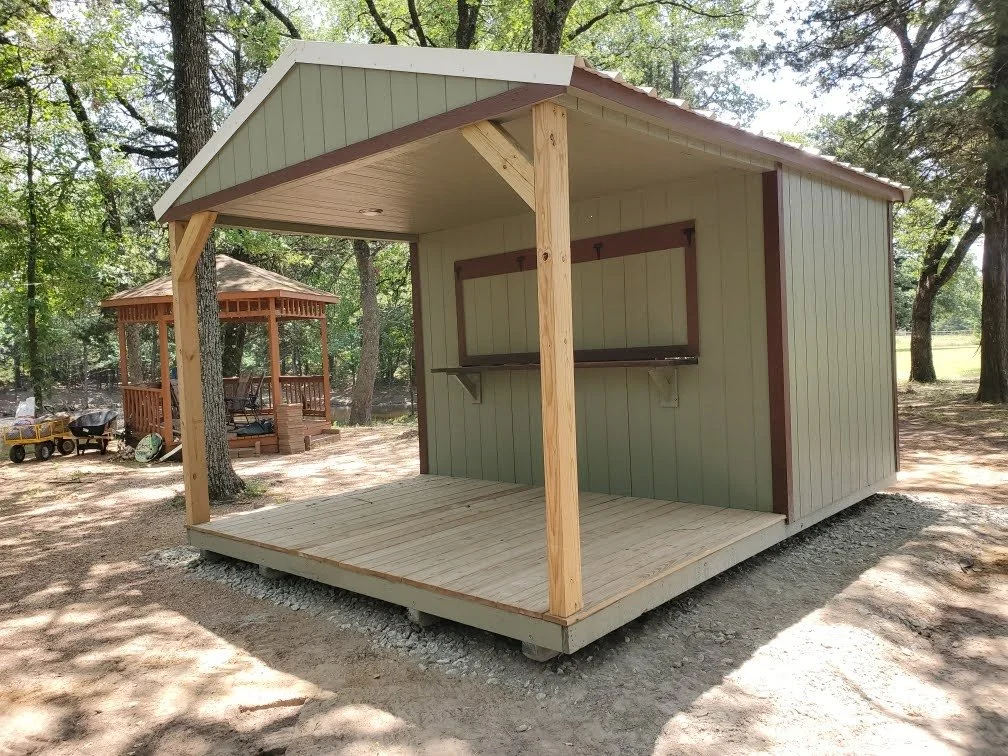 A small backyard shed with a porch and overhang, located in a wooded area with trees and another gazebo in the background.