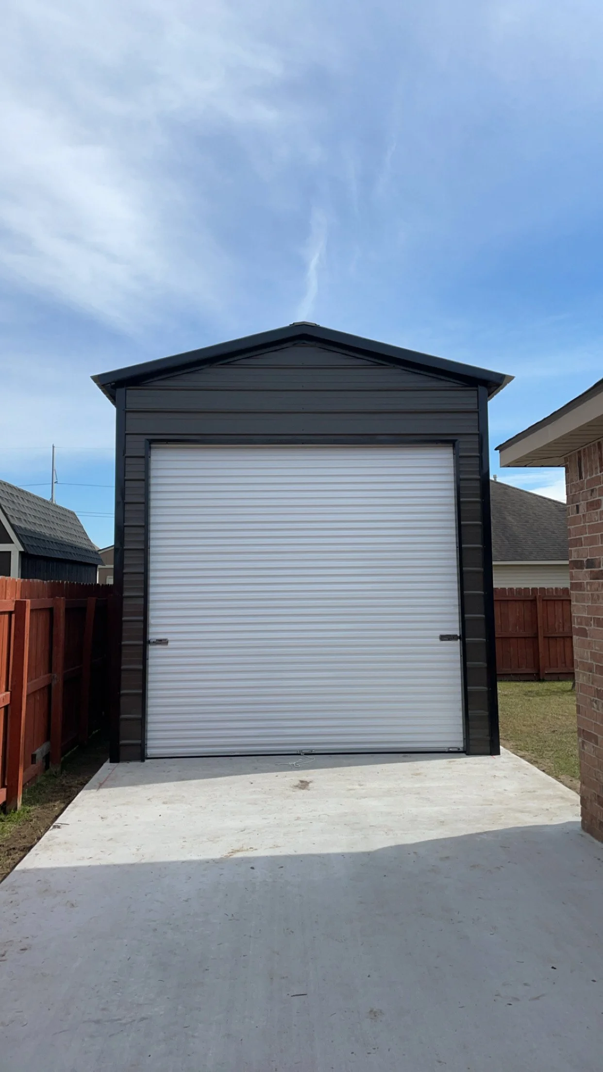 A black shed with a white roll-up garage door situated on a concrete slab, with a wooden fence on one side and brick house on the other, under a partly cloudy sky.