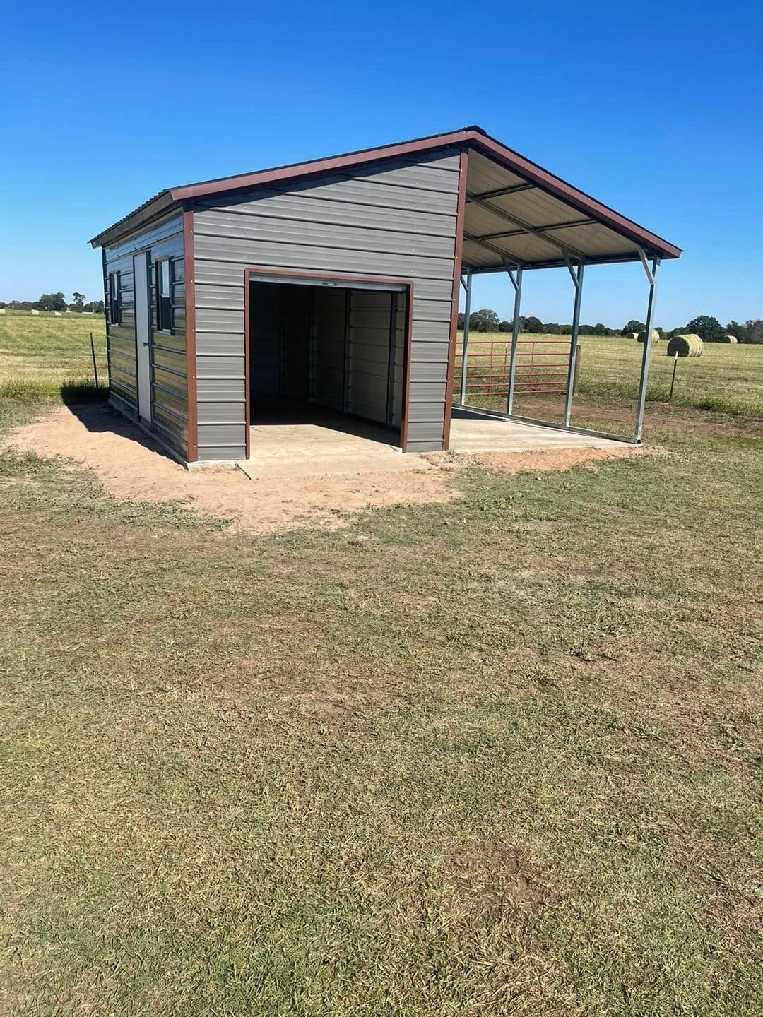 A metal shed with a partially open side, placed on a dirt patch in a grassy field, under a clear blue sky, with hay bales in the background.