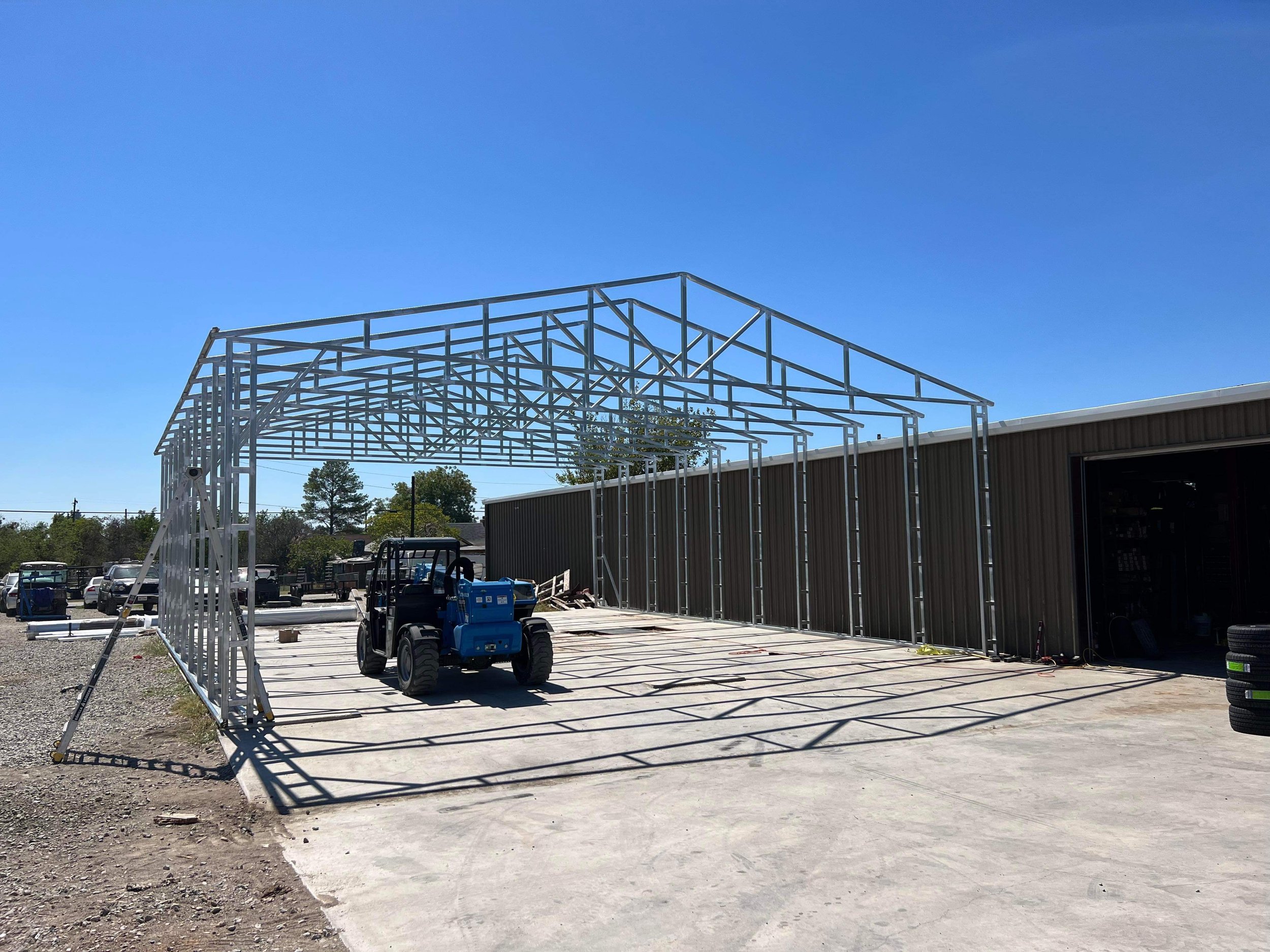 Metal frame structure under construction on a concrete lot with a forklift and tires, next to a brown building, under a clear blue sky.
